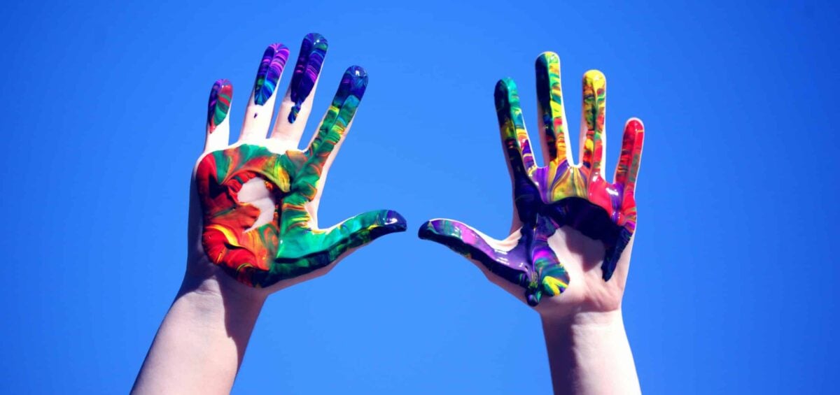 A person holding their hands covered in paint of many colors up against the sky.