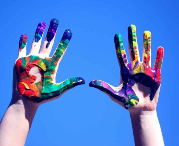 A person holding their hands covered in paint of many colors up against the sky.